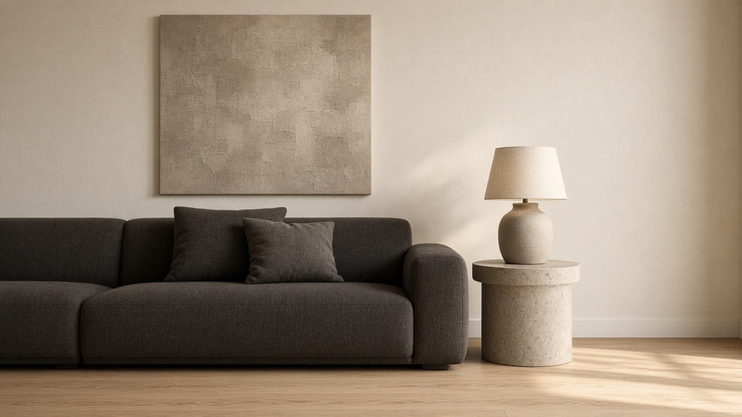 A charcoal wool sofa beneath a textured neutral artwork, with a stone side table and a ceramic lamp.
